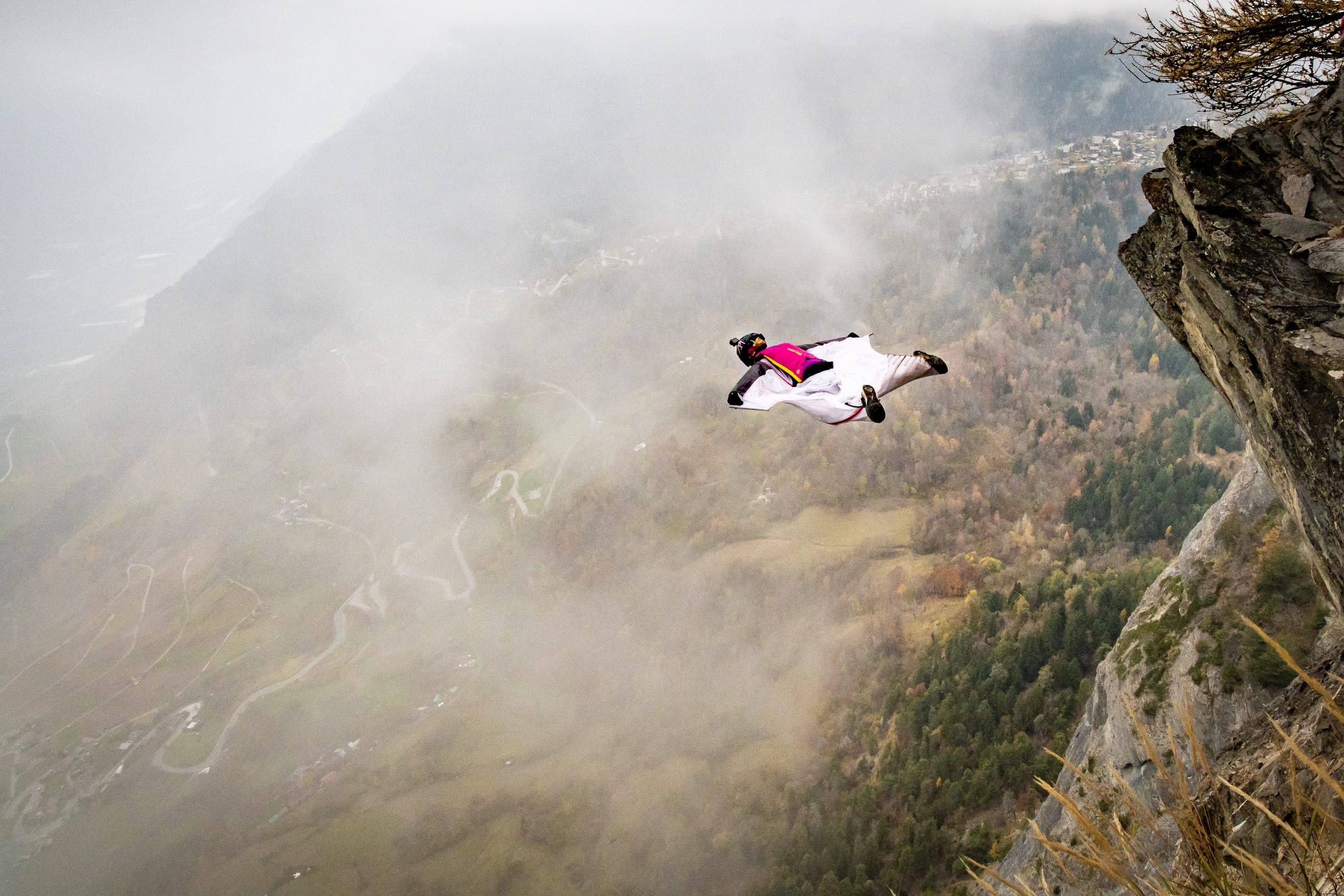 Géraldine Fasnacht en vol en wingsuit au-dessus d’une vallée verte, un lac turquoise et des sommets enneigés en contrebas.