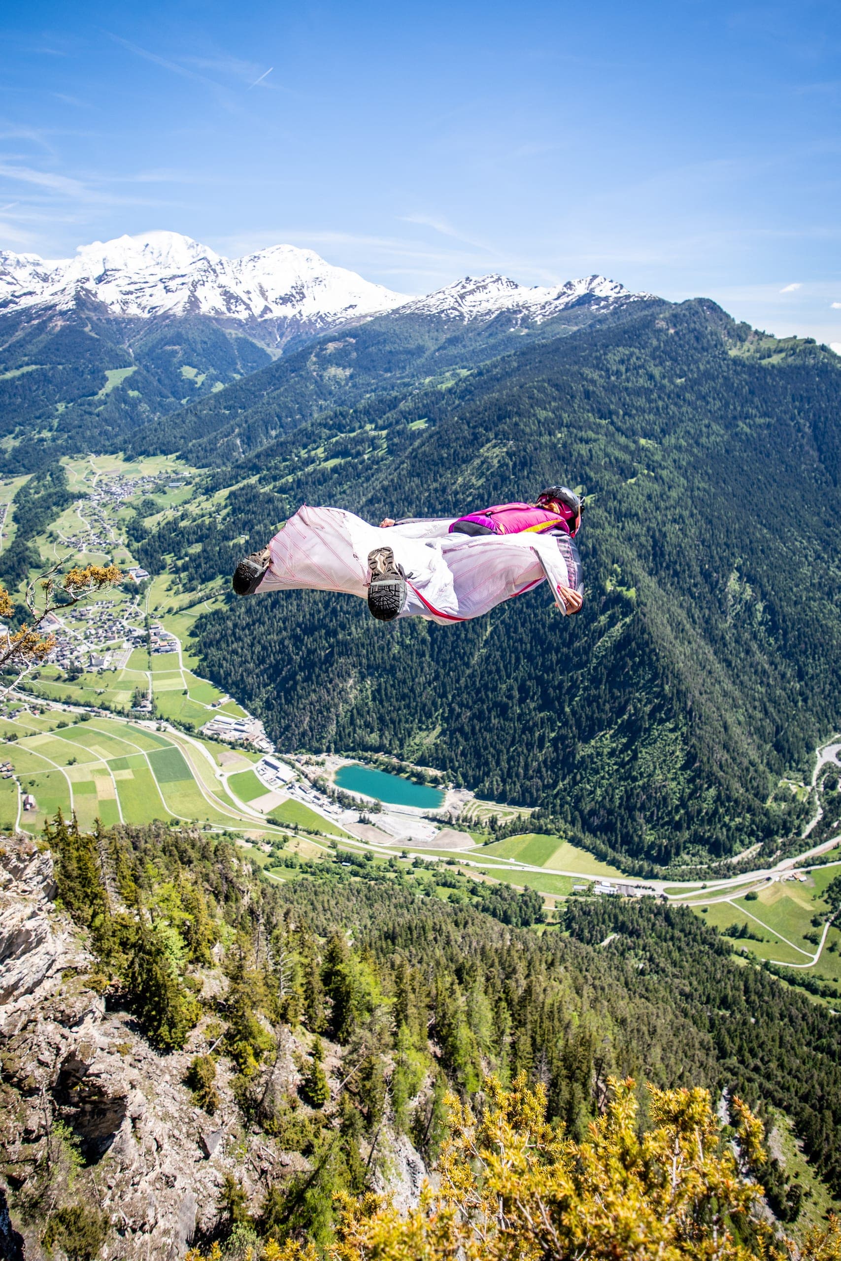 Géraldine Fasnacht en vol en wingsuit au-dessus d’une vallée verte, un lac turquoise et des sommets enneigés en contrebas.